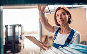 Frau mit blauer Latzhose und weißem Shirt hält Klemmbrett, greift im Lager nach Regal; unscharfer Gabelstapler im Hintergrund. Symbol für Inventur.
