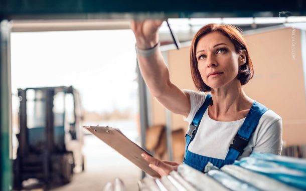 Frau mit blauer Latzhose und weißem Shirt hält Klemmbrett, greift im Lager nach Regal; unscharfer Gabelstapler im Hintergrund. Symbol für Inventur.